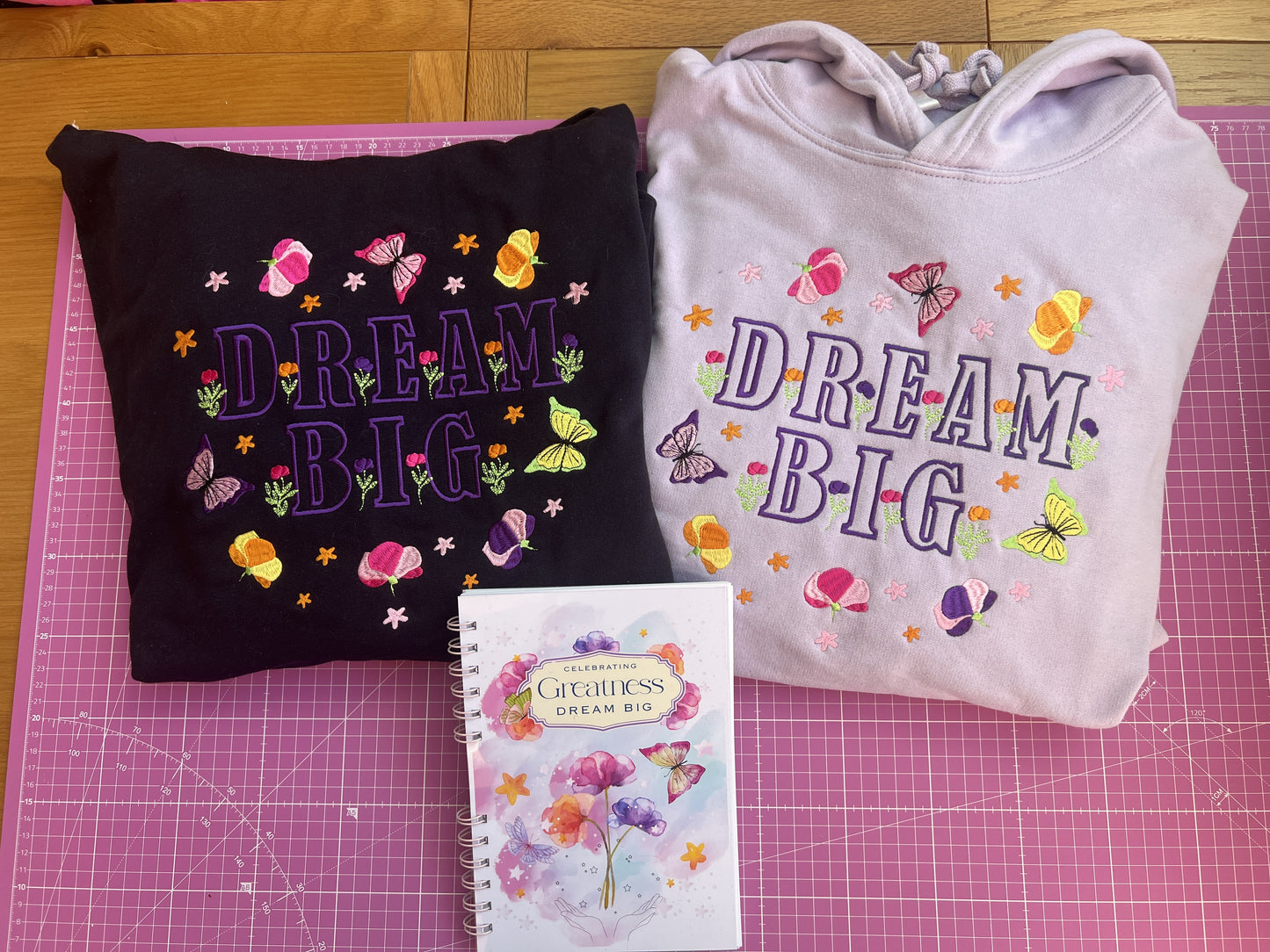 Black and Lilac sweatshirts with 'Dream Big' text on a pink cutting mat.