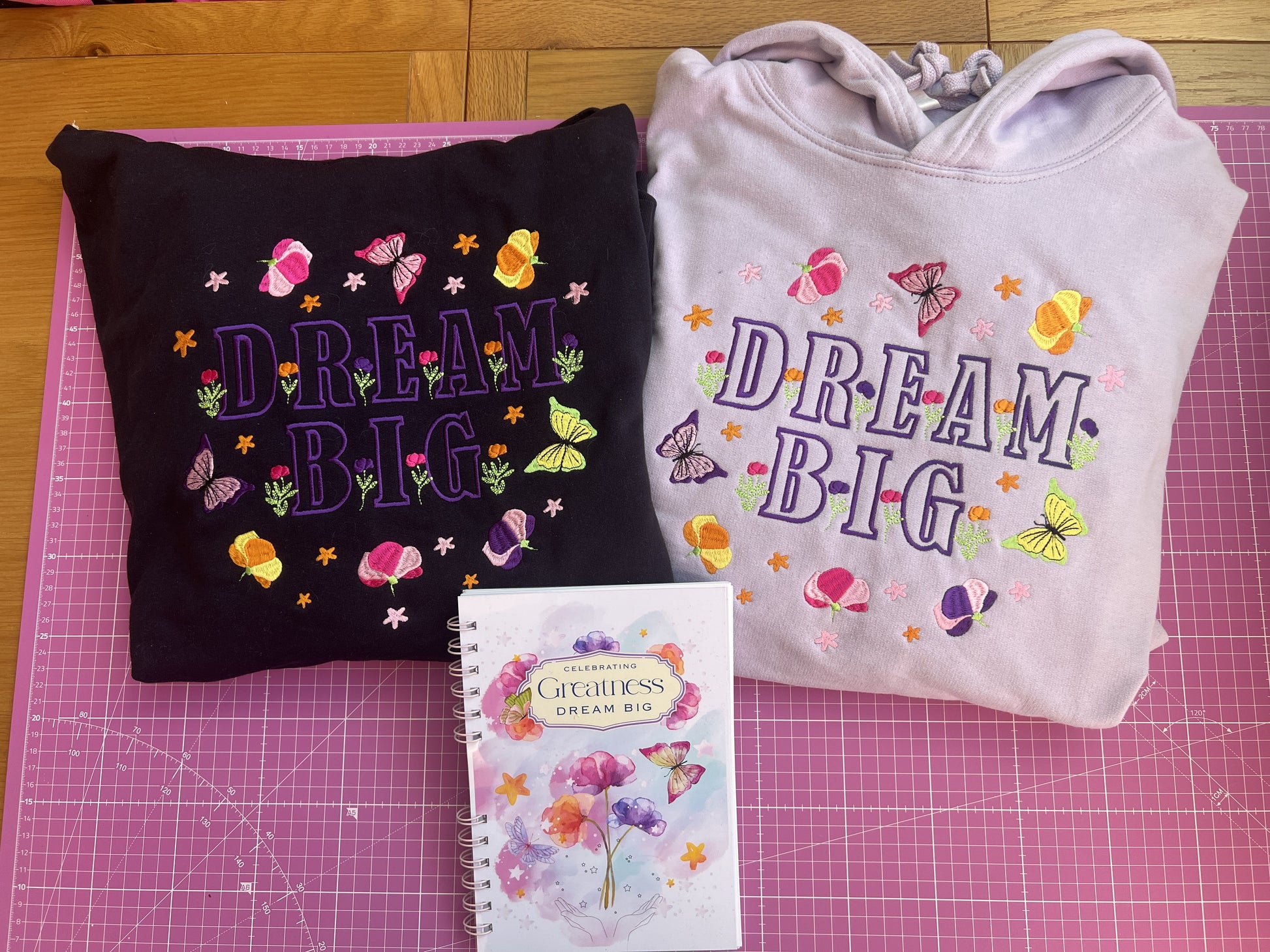 Black and Lilac sweatshirts with 'Dream Big' text on a pink cutting mat.