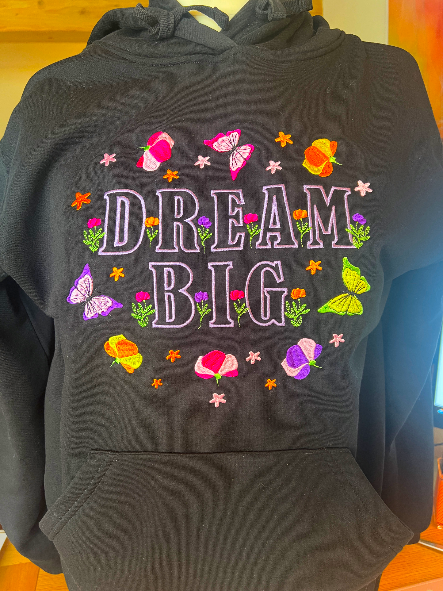 A black pullover hoodie featuring a colorful embroidered graphic on the front with the words 'DREAM BIG' surrounded by flowers, butterflies, and stars.