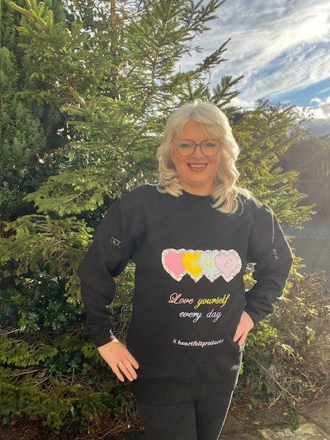 Person wearing a black sweatshirt with colorful hearts and text, standing outdoors.